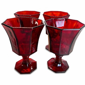 May include: A set of four deep red glass goblets featuring a faceted, octagonal bowl design and a matching hexagonal base. The translucent red glassware has a smooth, reflective surface and a distinct geometric shape with a sturdy stem.
