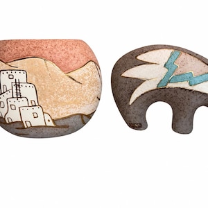 May include: A round ceramic pot featuring a painted desert landscape with white adobe structures and a separate dark gray ceramic bear figurine with a white and turquoise lightning bolt design on its side, both with textured, speckled surfaces.