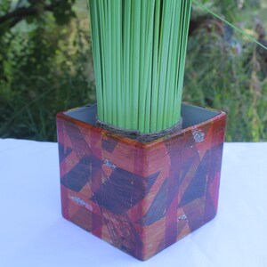 Peut inclure: Un cache-pot carré à la finition brillante orné d'un motif géométrique composé de coups de pinceau superposés rouge foncé, marron et orange. Le pot contient un bouquet dense de tiges d'herbe décoratives vert citron vif.