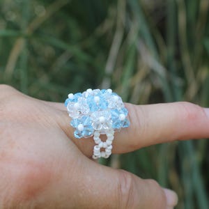 Op de afbeelding: Een kralenring met een cluster van lichtblauwe en doorzichtige gefacetteerde kristalkralen in een bloemmotief, geplaatst op een band gemaakt van kleine witte zaadkralen. Het ontwerp zorgt voor een getextureerd en meerdimensionaal uiterlijk.