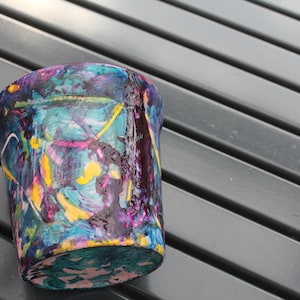 Peut inclure: Un petit pot de fleurs cylindrique orné d'un motif de peinture abstrait et multicolore. La surface est recouverte de traînées et d'éclaboussures vibrantes de peinture bleue, violette, jaune et noire, créant un aspect texturé et superposé.