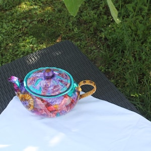 Puede incluir: Una tetera redonda con un patrón abstracto vibrante en tonos morado, rosa y naranja. La tapa y el borde están acentuados con un borde turquesa brillante, y el asa presenta un patrón distintivo de manchas en color marrón y negro.