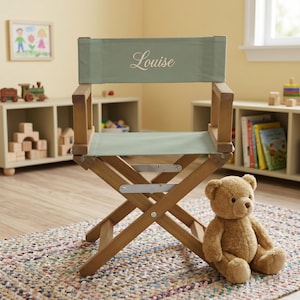 May include: A wooden director's chair with a sage green fabric seat and backrest. The backrest features the name Louise embroidered in white script. The chair has a folding frame with metal hardware and is positioned next to a brown teddy bear.