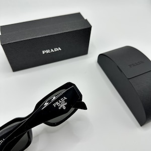 Op de afbeelding: Zwarte Prada zonnebril met het Prada logo op de poot. De zonnebril ligt naast een zwarte Prada etui en een zwarte Prada doos. De zonnebril heeft een rechthoekig montuur.