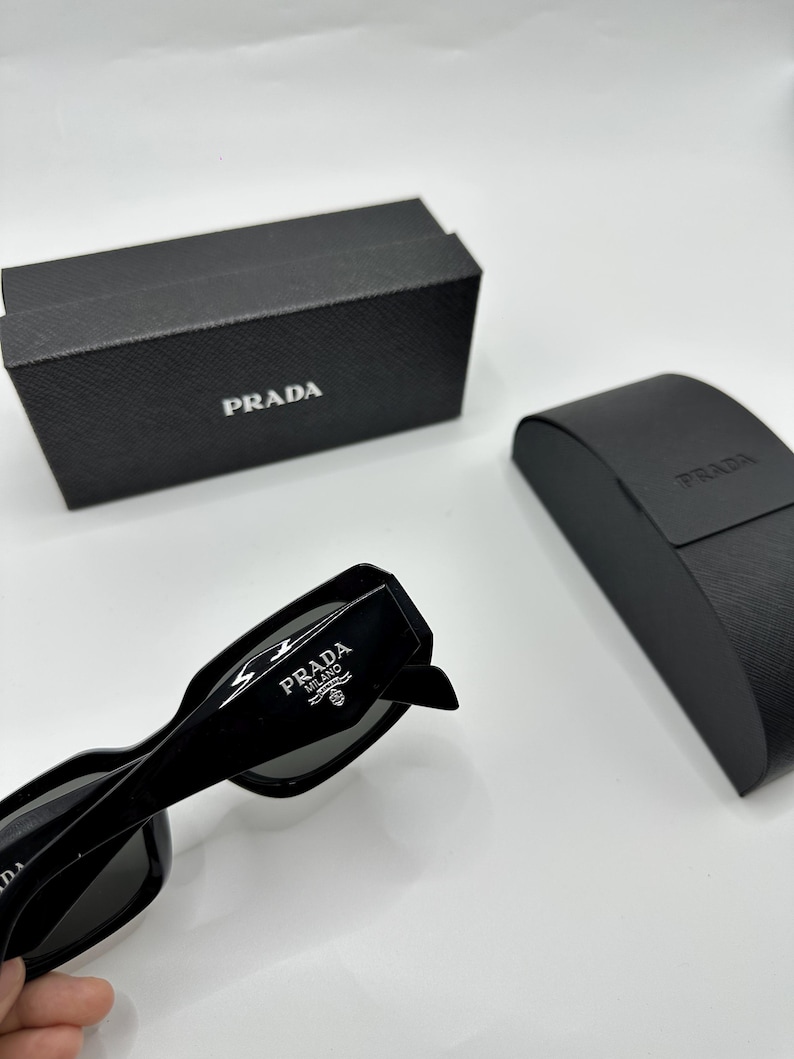 Op de afbeelding: Zwarte Prada zonnebril met het Prada logo op de poot. De zonnebril ligt naast een zwarte Prada etui en een zwarte Prada doos. De zonnebril heeft een rechthoekig montuur.