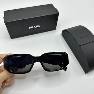 Op de afbeelding: Zwarte Prada zonnebril met een rechthoekig montuur en donkere glazen. De zonnebril wordt getoond met een zwarte Prada etui en doos, beide met het Prada logo.