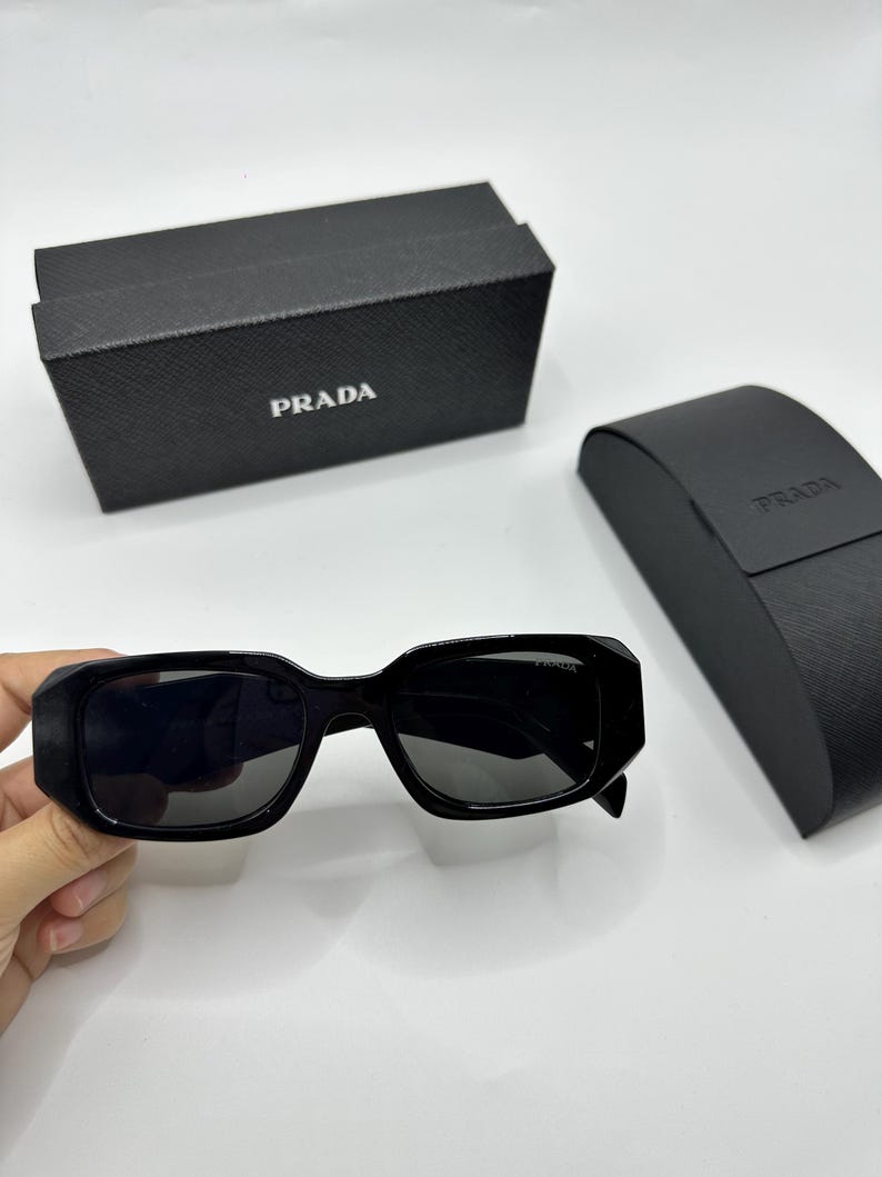 Op de afbeelding: Zwarte Prada zonnebril met een rechthoekig montuur en donkere glazen. De zonnebril wordt getoond met een zwarte Prada etui en doos, beide met het Prada logo.