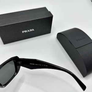 Op de afbeelding: Zwarte Prada zonnebril met bijpassende etui en doos. De zonnebril heeft donkere glazen en een strak, modern design. Het Prada-logo is zichtbaar op de etui, doos en de brilpoot. De doos en etui zijn zwart.