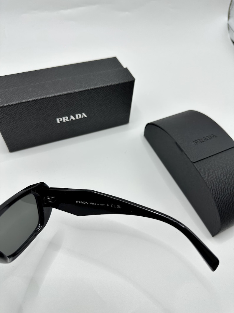 Op de afbeelding: Zwarte Prada zonnebril met bijpassende etui en doos. De zonnebril heeft donkere glazen en een strak, modern design. Het Prada-logo is zichtbaar op de etui, doos en de brilpoot. De doos en etui zijn zwart.