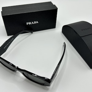 Op de afbeelding: Zwarte Prada zonnebril met bijpassende etui en doos. De zonnebril heeft een rechthoekig montuur en donkere glazen. De doos en etui zijn zwart met het Prada-logo in wit. Op een witte ondergrond.