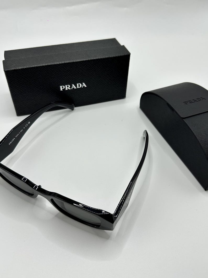 Op de afbeelding: Zwarte Prada zonnebril met bijpassende etui en doos. De zonnebril heeft een rechthoekig montuur en donkere glazen. De doos en etui zijn zwart met het Prada-logo in wit. Op een witte ondergrond.
