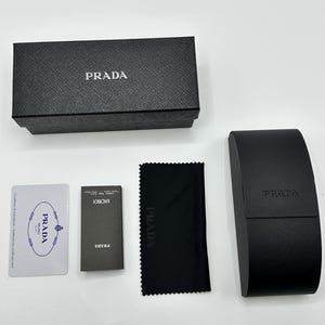 Op de afbeelding: Een zwarte Prada brillenkoker, doos, schoonmaakdoek en papierwerk worden getoond op een witte ondergrond. Het Prada-logo is zichtbaar op de doos, koker en doek. De doos en koker hebben een getextureerde afwerking.