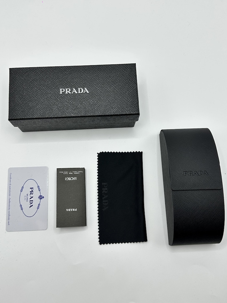 Op de afbeelding: Een zwarte Prada brillenkoker, doos, schoonmaakdoek en papierwerk worden getoond op een witte ondergrond. Het Prada-logo is zichtbaar op de doos, koker en doek. De doos en koker hebben een getextureerde afwerking.