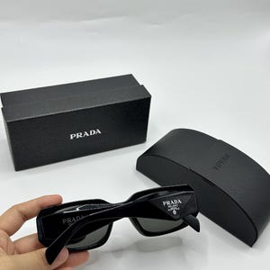 Op de afbeelding: Zwarte Prada zonnebril met bijpassende etui en doos. De zonnebril heeft een rechthoekig montuur en donkere glazen. Het Prada-logo is zichtbaar op de pootjes en de doos. De etui is ook zwart en heeft het Prada-logo.