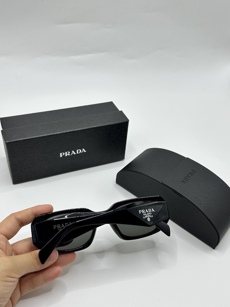 Op de afbeelding: Zwarte Prada zonnebril met bijpassende etui en doos. De zonnebril heeft een rechthoekig montuur en donkere glazen. Het Prada-logo is zichtbaar op de pootjes en de doos. De etui is ook zwart en heeft het Prada-logo.