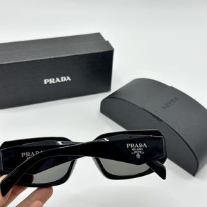 Op de afbeelding: Zwarte rechthoekige zonnebril met grijze glazen. De zonnebril heeft het witte "PRADA MILANO" logo op de poot. Een zwarte etui en doos met het "PRADA" logo zijn ook zichtbaar.
