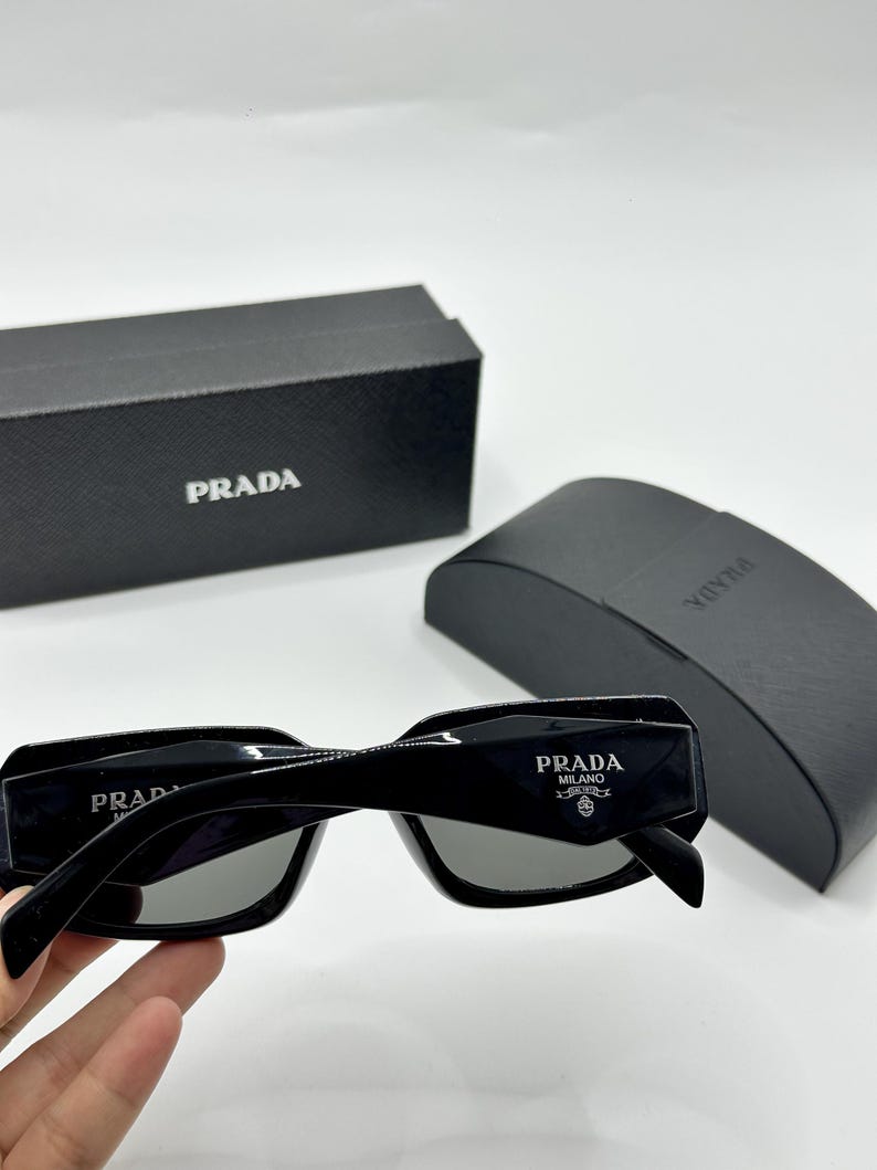 Op de afbeelding: Zwarte rechthoekige zonnebril met grijze glazen. De zonnebril heeft het witte "PRADA MILANO" logo op de poot. Een zwarte etui en doos met het "PRADA" logo zijn ook zichtbaar.