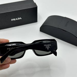 Op de afbeelding: Zwarte Prada zonnebril met grijze glazen, met het Prada Milano logo op de poot. De zonnebril wordt geleverd met een zwarte Prada etui en doos, beide met het Prada logo.