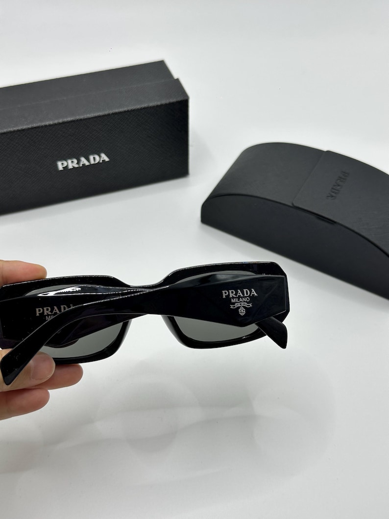 Op de afbeelding: Zwarte Prada zonnebril met grijze glazen, met het Prada Milano logo op de poot. De zonnebril wordt geleverd met een zwarte Prada etui en doos, beide met het Prada logo.