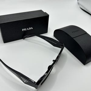 Op de afbeelding: Zwarte Prada zonnebril met bijpassende etui en doos. De zonnebril heeft een rechthoekig montuur en het Prada-logo is zichtbaar op de pootjes en de doos. De etui is zwart en heeft het Prada-logo.