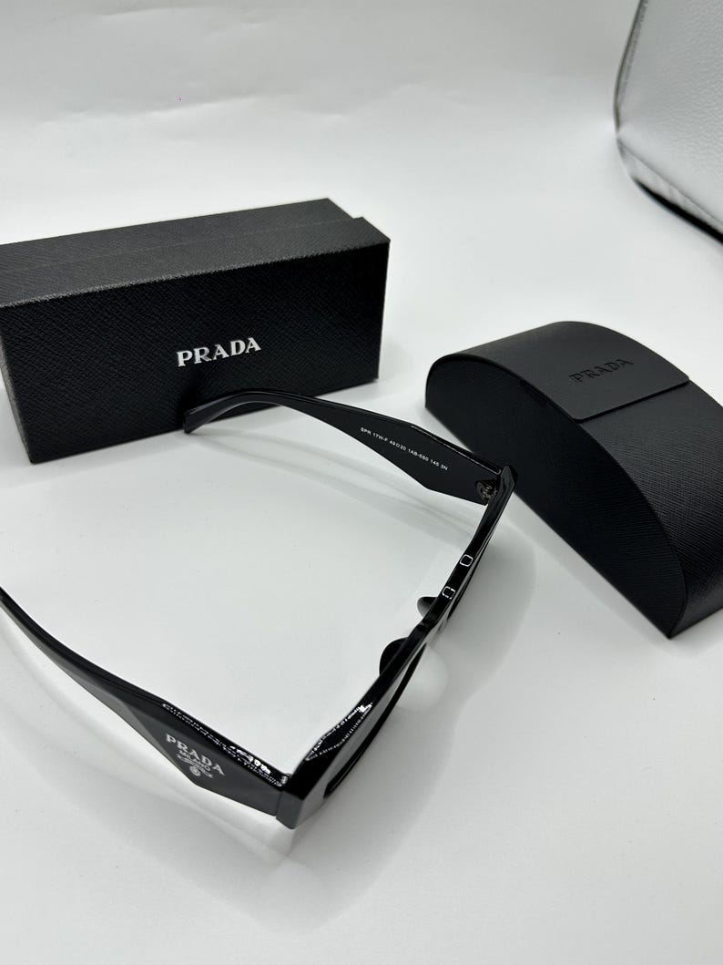 Op de afbeelding: Zwarte Prada zonnebril met bijpassende etui en doos. De zonnebril heeft een rechthoekig montuur en het Prada-logo is zichtbaar op de pootjes en de doos. De etui is zwart en heeft het Prada-logo.