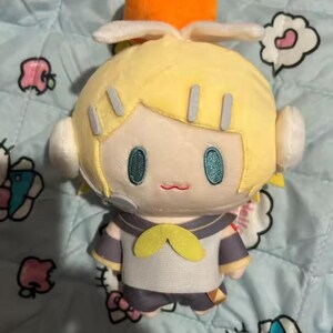 May include: A plush doll with yellow hair, teal eyes, and a small red mouth. The doll wears a sailor-style outfit with a yellow bow tie and a small orange fruit on its head. The doll is on a light blue patterned surface.