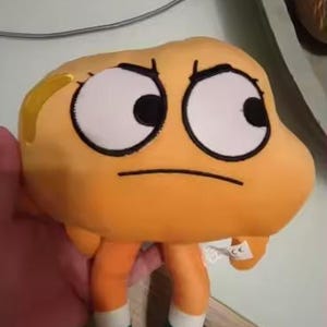 May include: Orange plush toy of a cartoon character with large white eyes, a black frown, and orange legs and arms. The character is wearing green and white shoes. A small panda plush toy is in the background.