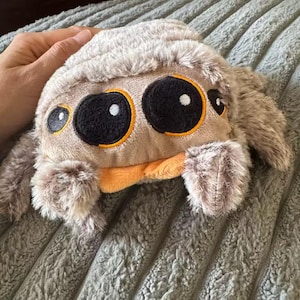May include: A plush toy spider with large, round eyes outlined in orange and black. The spider has a tan body with fluffy, gray fur and orange mouth. The toy is designed to be soft and cuddly.