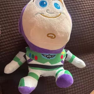 Puede incluir: Un peluche de Buzz Lightyear, con una cabeza blanca, ojos azules y un casco morado. El juguete tiene un traje espacial verde y blanco con detalles morados. Est&aacute; sobre una superficie marr&oacute;n.