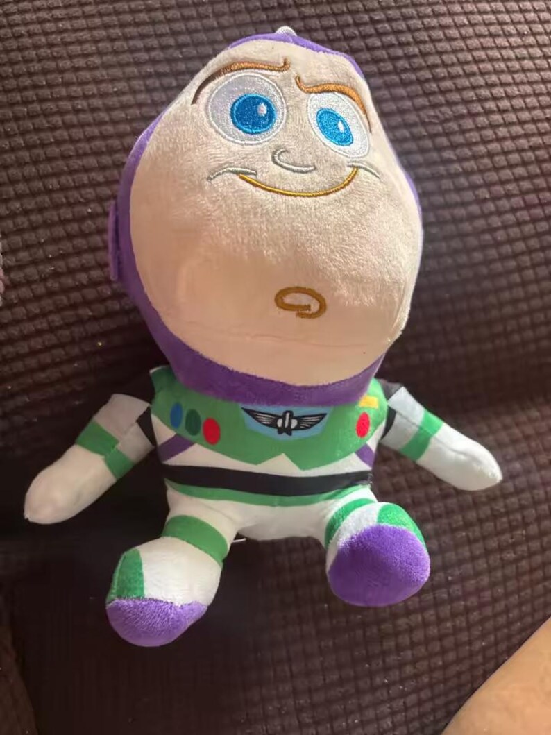 Puede incluir: Un peluche de Buzz Lightyear, con una cabeza blanca, ojos azules y un casco morado. El juguete tiene un traje espacial verde y blanco con detalles morados. Est&aacute; sobre una superficie marr&oacute;n.