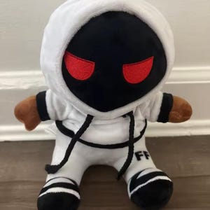 May include: A plush doll in a white hooded sweatshirt and matching pants with black stripes. The doll has a black face with red eyes and brown arms. The letters "FF" are printed on the left leg of the doll.