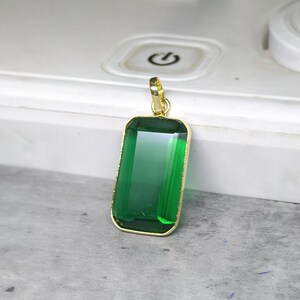 May include: A rectangular pendant with a large, emerald-green gemstone set in a gold-colored frame. The pendant has a simple, elegant design with a gold bail for attaching to a chain. The gemstone is faceted, reflecting light.