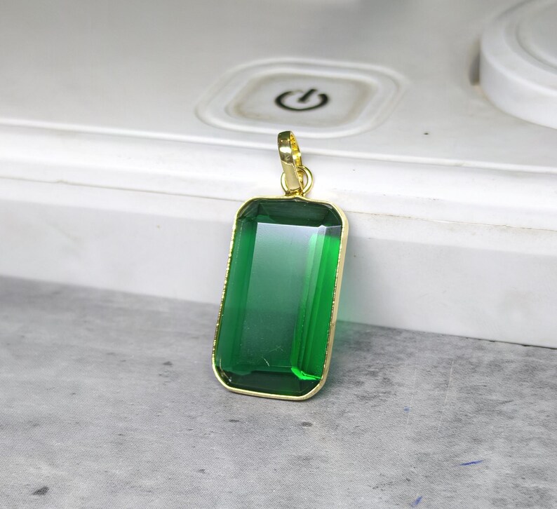 May include: A rectangular pendant with a large, emerald-green gemstone set in a gold-colored frame. The pendant has a simple, elegant design with a gold bail for attaching to a chain. The gemstone is faceted, reflecting light.