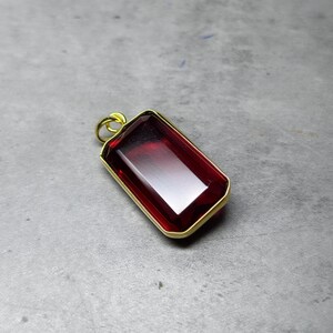 May include: A rectangular pendant featuring a deep red gemstone, set within a gold-coloured frame. The polished gemstone reflects light. The pendant has a small loop at the top for a chain, suitable for necklaces.