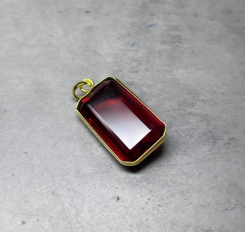 May include: A rectangular pendant featuring a deep red gemstone, set within a gold-coloured frame. The polished gemstone reflects light. The pendant has a small loop at the top for a chain, suitable for necklaces.