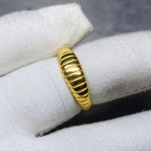 Könnte beinhalten: Nahaufnahme eines goldenen Rings mit geripptem Design. Der Ring hat eine helle, glänzende Goldfarbe und scheint aus Metall zu sein. Er liegt auf einer weißen Stoffoberfläche.