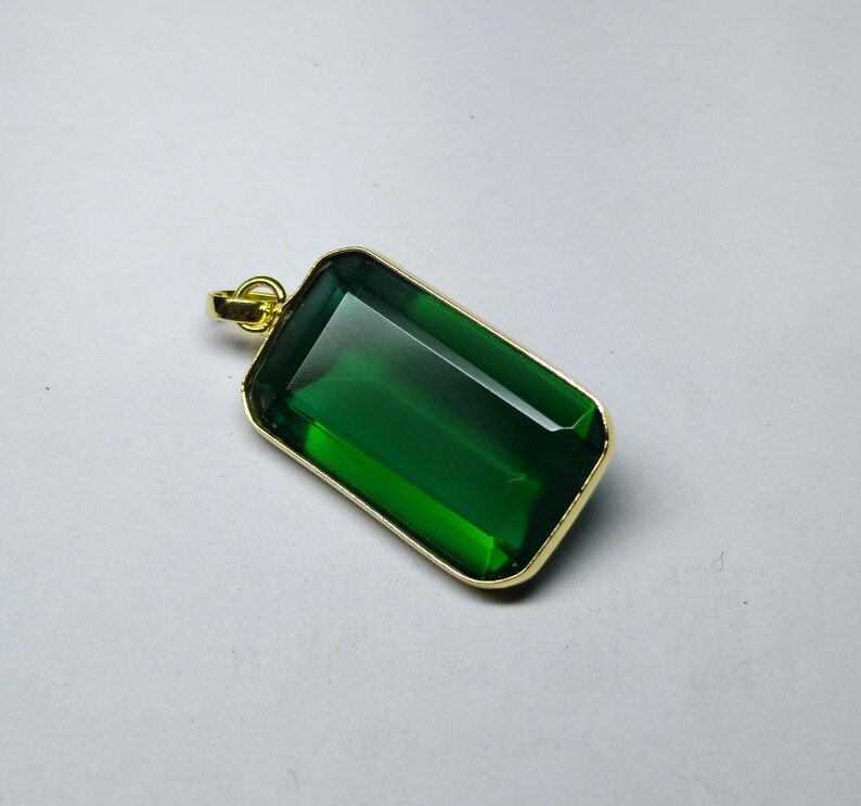 May include: A rectangular green gemstone pendant with a gold-colored frame and bail. The emerald-colored stone is set in a gold-tone metal. The pendant is a simple, elegant design.