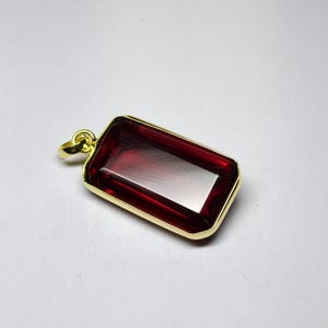 May include: A rectangular pendant featuring a deep red gemstone set within a gold-coloured frame. The gemstone has a polished, reflective surface. The pendant has a small loop at the top for a chain. Jewellery.