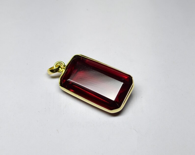 May include: A rectangular pendant featuring a deep red gemstone set within a gold-coloured frame. The gemstone has a polished, reflective surface. The pendant has a small loop at the top for a chain. Jewellery.