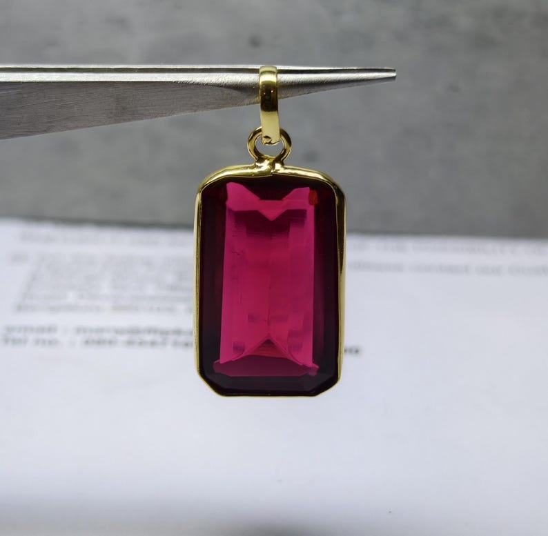 May include: A gold-coloured pendant featuring a rectangular, magenta gemstone. The gemstone is set within a gold frame and suspended from a gold bail. The pendant is held by tweezers, ready for a chain.