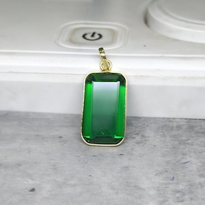 Puede incluir: Un colgante rectangular de piedra verde con un marco dorado. El colgante tiene un efecto degradado, que pasa de un verde más oscuro en la parte superior a un verde más claro en la parte inferior. El colgante está suspendido de una fianza dorada.
