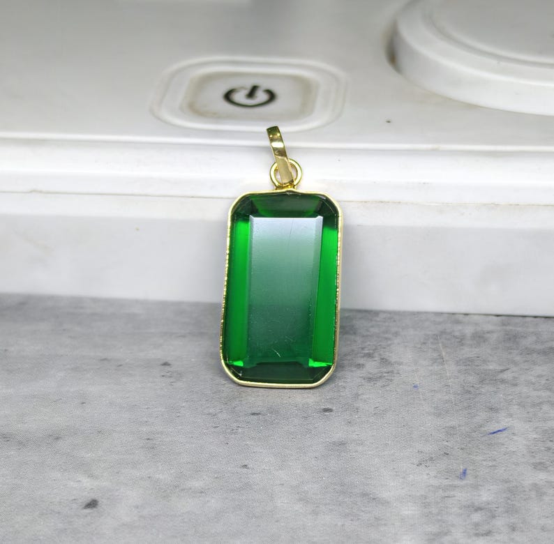 May include: A rectangular green gemstone pendant with a gold-colored frame. The pendant has a gradient effect, transitioning from a darker green at the top to a lighter green at the bottom. The pendant is suspended from a gold-colored bail.