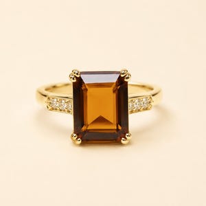 May include: A gold ring featuring a large, rectangular, emerald-cut amber-colored gemstone set in the center. The band is crafted from polished yellow gold and includes three small, clear, round-cut accent stones embedded on each side.