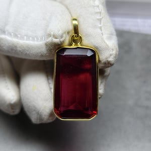 May include: A rectangular pendant featuring a deep red gemstone within a gold-coloured frame. The gemstone is faceted, and the pendant has a loop for a chain. The pendant is held by a gloved hand.