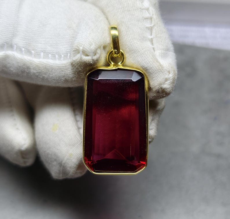 May include: A rectangular pendant featuring a deep red gemstone within a gold-coloured frame. The gemstone is faceted, and the pendant has a loop for a chain. The pendant is held by a gloved hand.