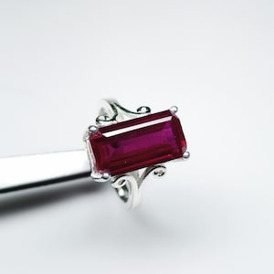 Puede incluir: Un anillo de plata con una gran gema rectangular de color rojo intenso. El anillo tiene un diseño ornamental y arremolinado alrededor de la gema, con pequeños elementos decorativos en las esquinas.