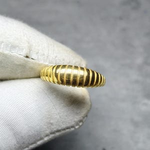 Könnte beinhalten: Ein goldener Ring mit geripptem Design. Der Ring hat eine leuchtend gelbe Farbe und scheint aus Metall zu sein. Der Ring wird in einem weißen Tuch gehalten. Der Ring ist ein Schmuckstück.