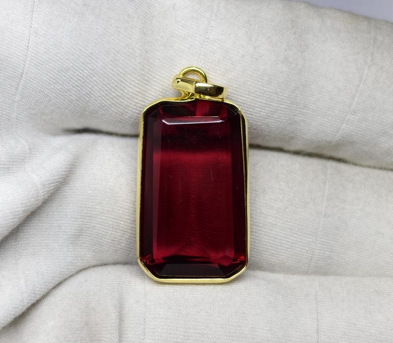 May include: A rectangular pendant featuring a deep red gemstone set within a gold-coloured frame. The gemstone is a rich, transparent red. The pendant has a loop at the top for a chain. The background is a white, textured fabric.