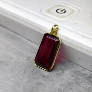 May include: A rectangular pendant featuring a deep red gemstone within a gold-coloured frame. The gemstone's faceted surface reflects light. The pendant's design is simple and elegant, suitable for various occasions. The pendant is likely for a necklace.