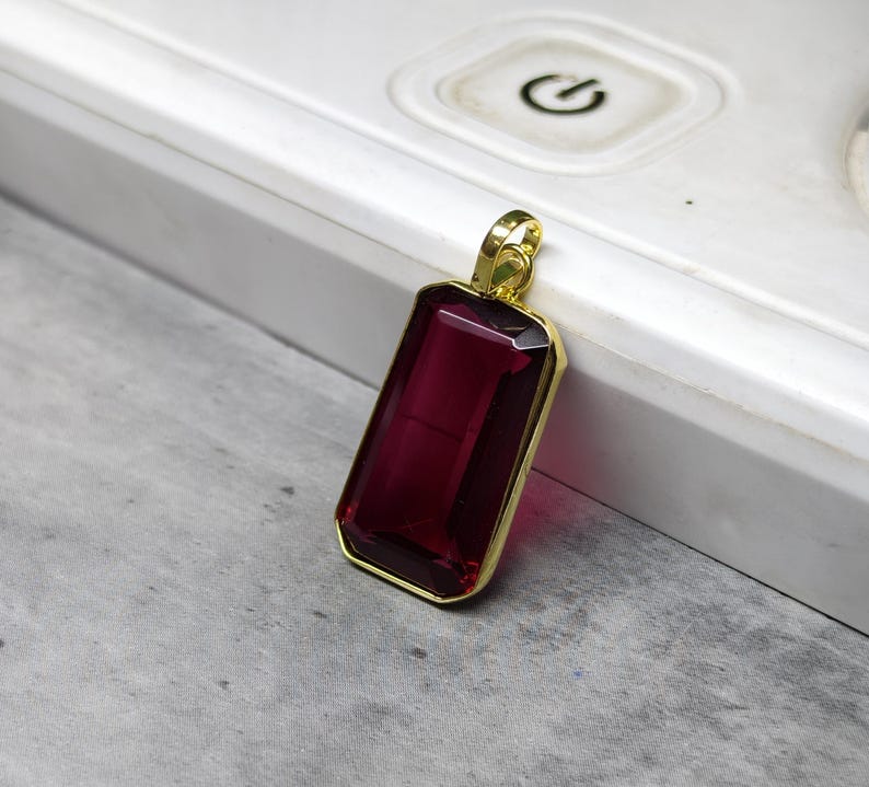 May include: A rectangular pendant featuring a deep red gemstone within a gold-coloured frame. The gemstone's faceted surface reflects light. The pendant's design is simple and elegant, suitable for various occasions. The pendant is likely for a necklace.